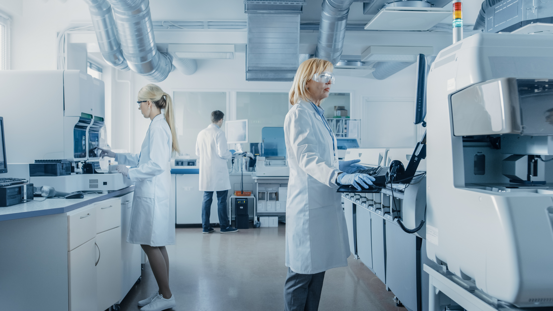 Two women and a man wearing white coats and protective goggles stand in a laboratory.