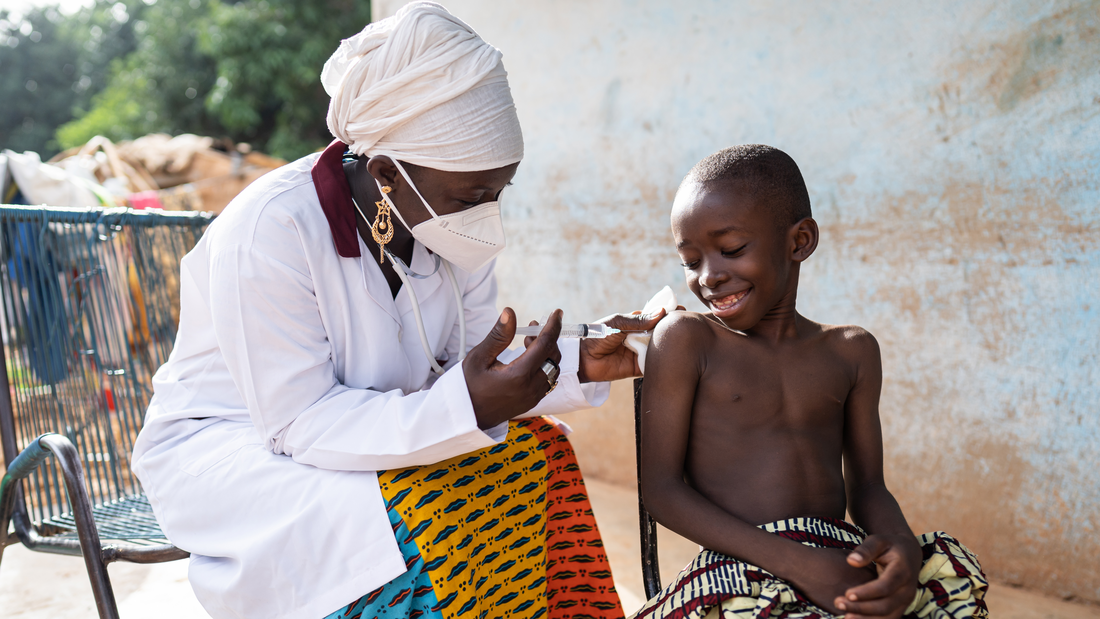 A woman in a white coat sticks a syringe into the arm of a laughing child.